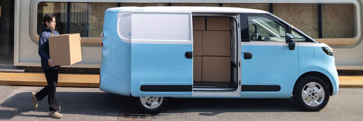 Ein junger Mann trägt einen Karton zu einem blauen Lieferwagen mit offenem Heck, das mit Kisten beladen ist.