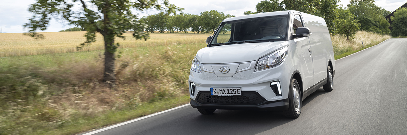 Ein weißer Lieferwagen fährt auf einer Landstraße, umgeben von grünen Bäumen und Feldern.