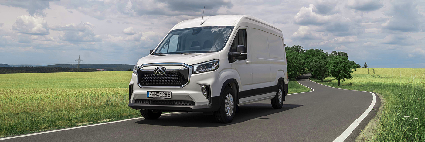 Weißer Transporter fährt auf einer Kurvenstraße durch eine grüne Landschaft unter bewölktem Himmel.
