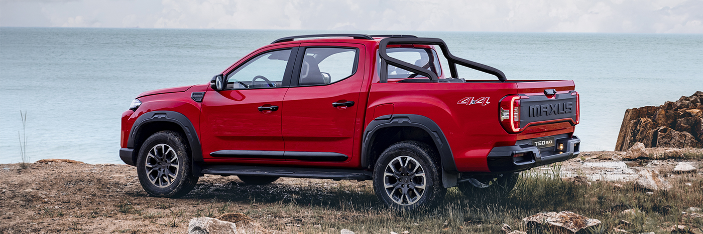 Roter Pickup-Truck am Strand mit Aussicht auf das Meer, im Hintergrund Steine und eine bewölkte Himmel.